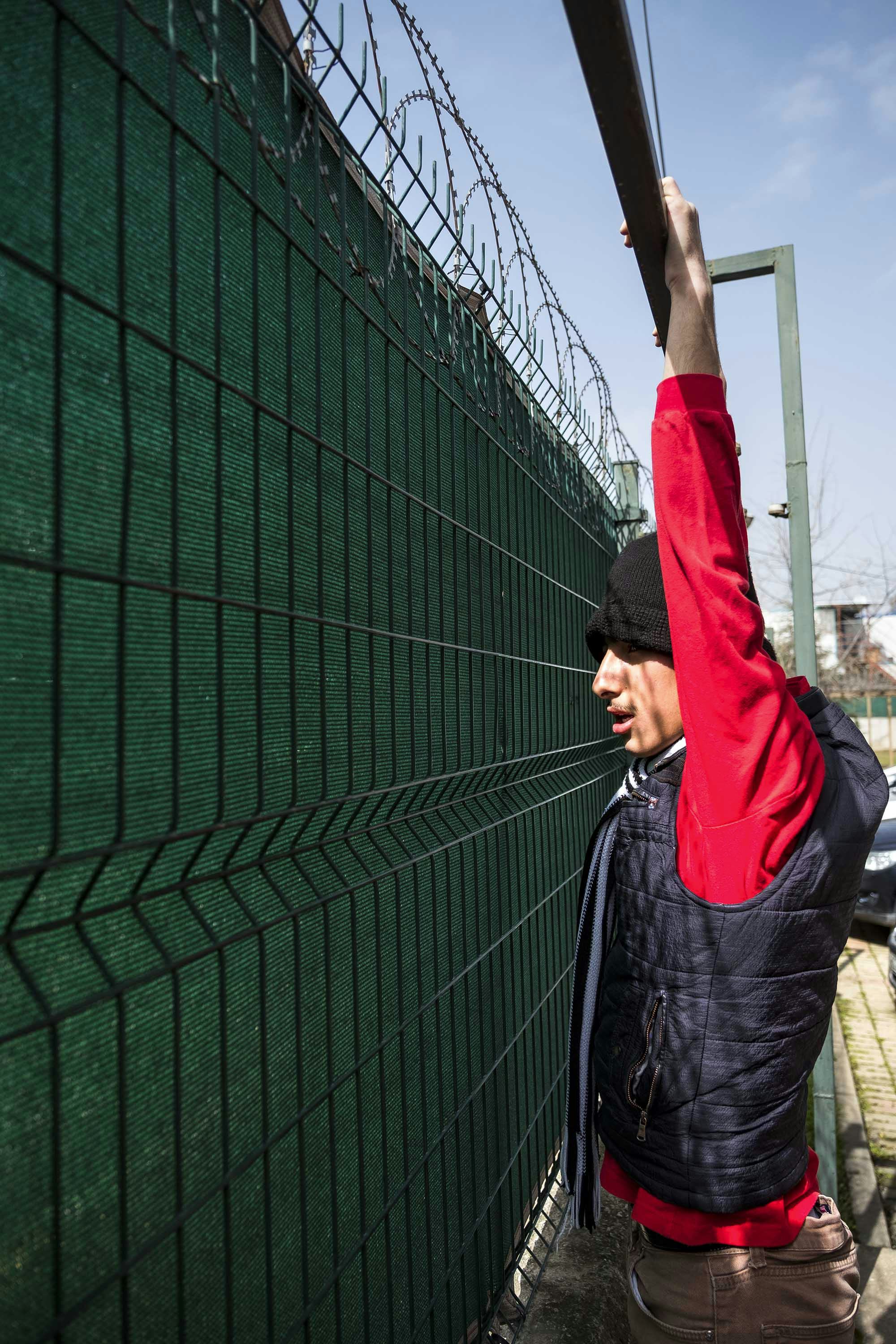 Photo of the boy in a red sweater hanging on a high, closed fence. © Oğulcan Arslan