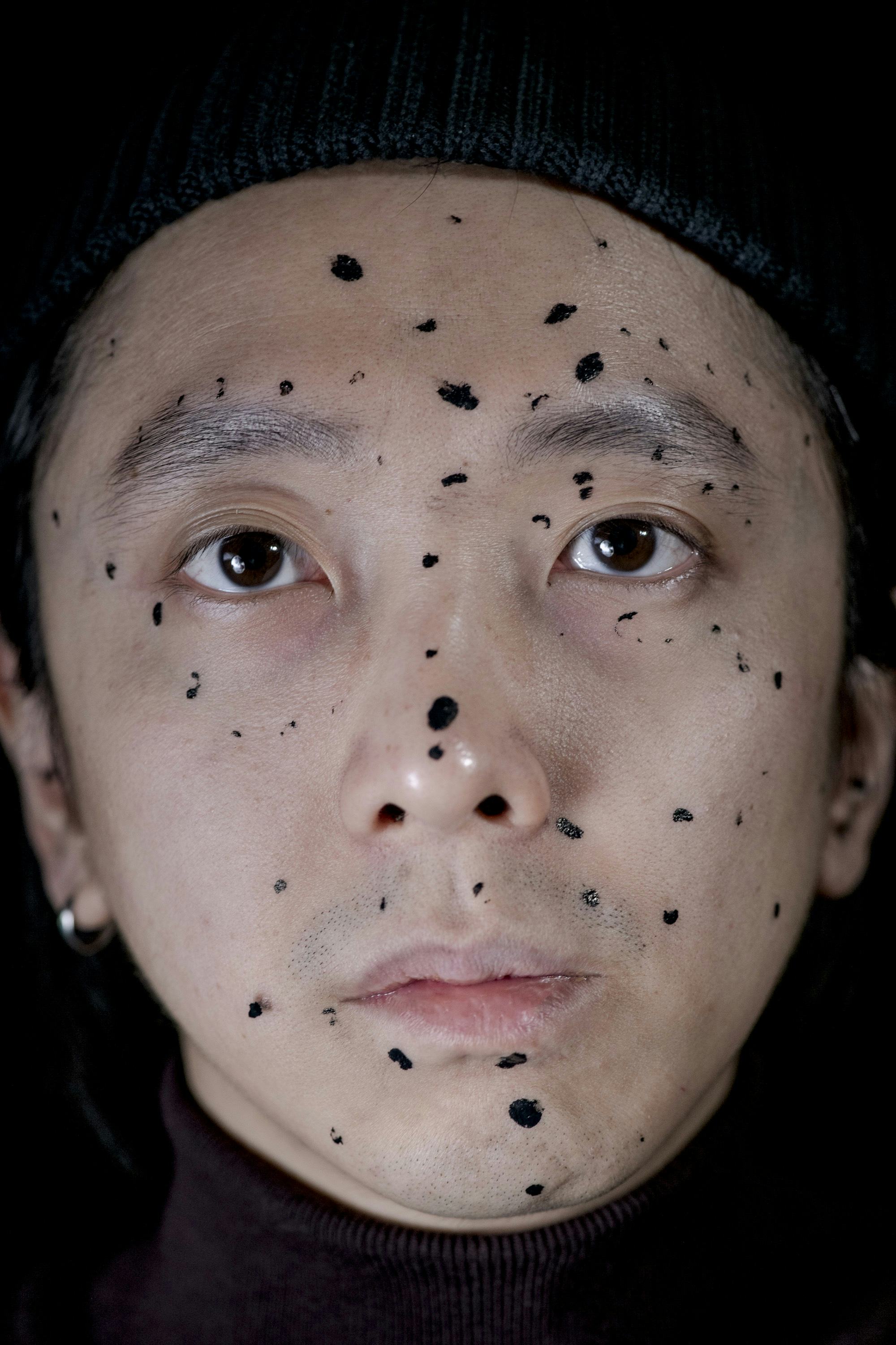 Frontal portrait of the artist's face, his face marked by black paint dots of different shapes and sizes© Sheung Yiu