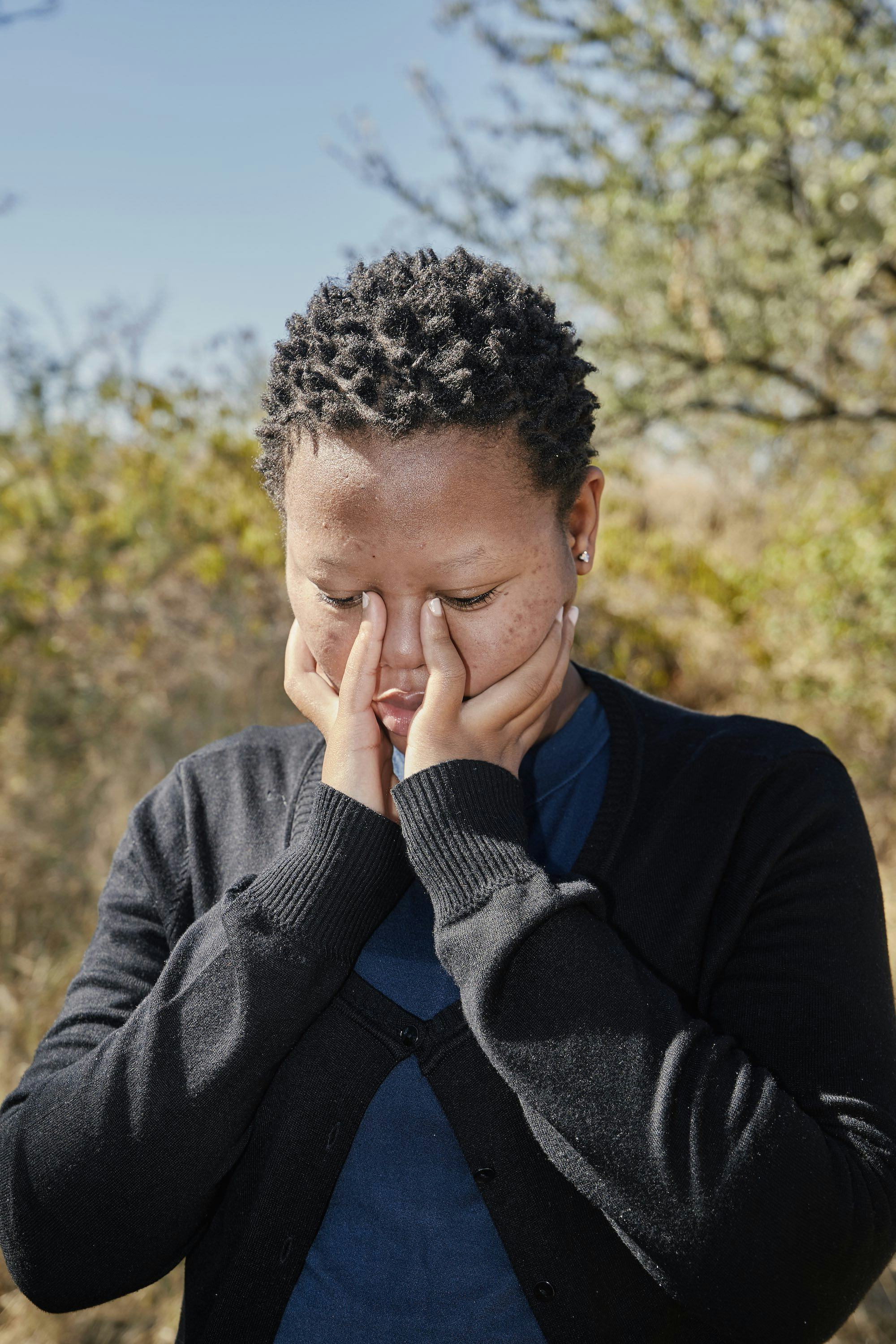Portrait of a young Black woman in a black cardigan, holding her face in her hands© Thero Makepe