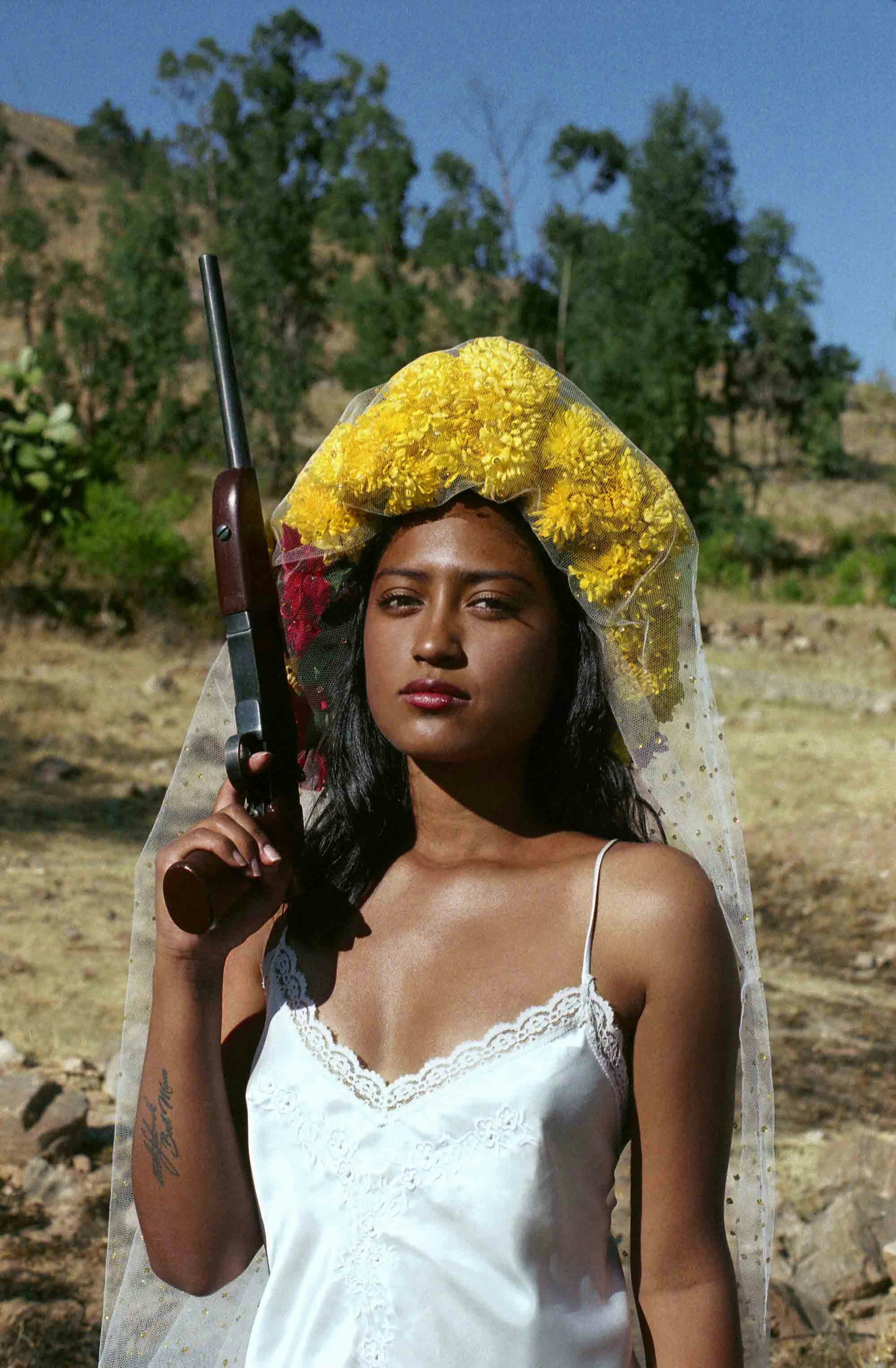 Portrait of a young woman outdoors, wearing a white neglige dress and a bright yellow flower crown, holding a rifle up in the air© Marisol Mendez