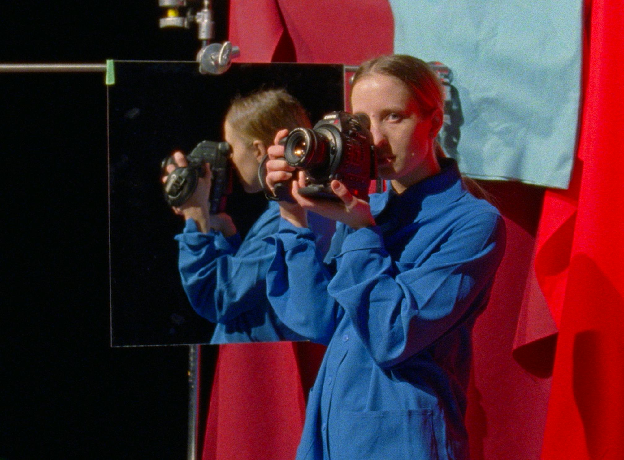 Self portrait of the artist Sara Cwynar, holding a camera in front of a mirror, in front of a red curtain.