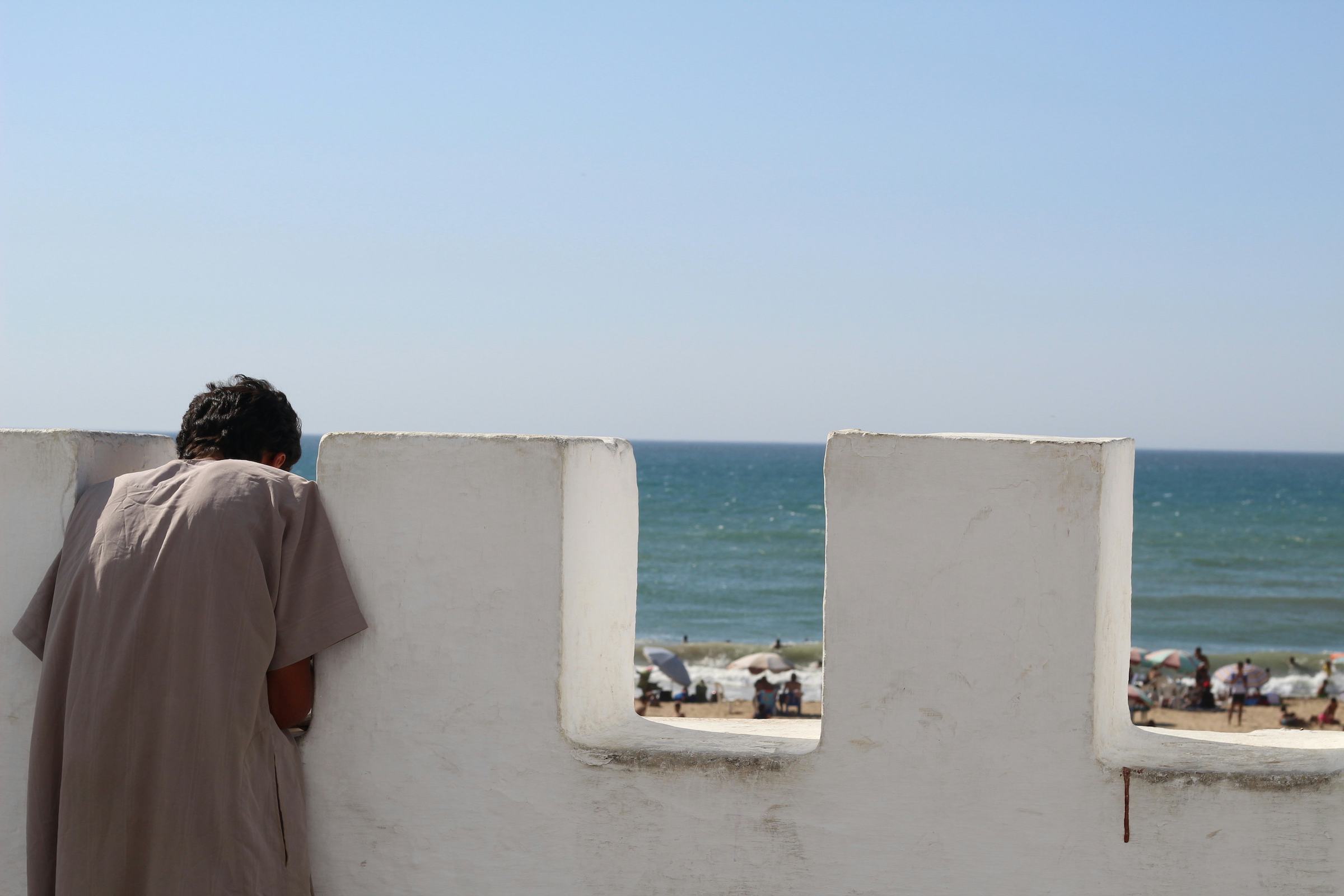 Photo of a young man from the back overlooking the sea through a white wall. © Dina Bousbaa