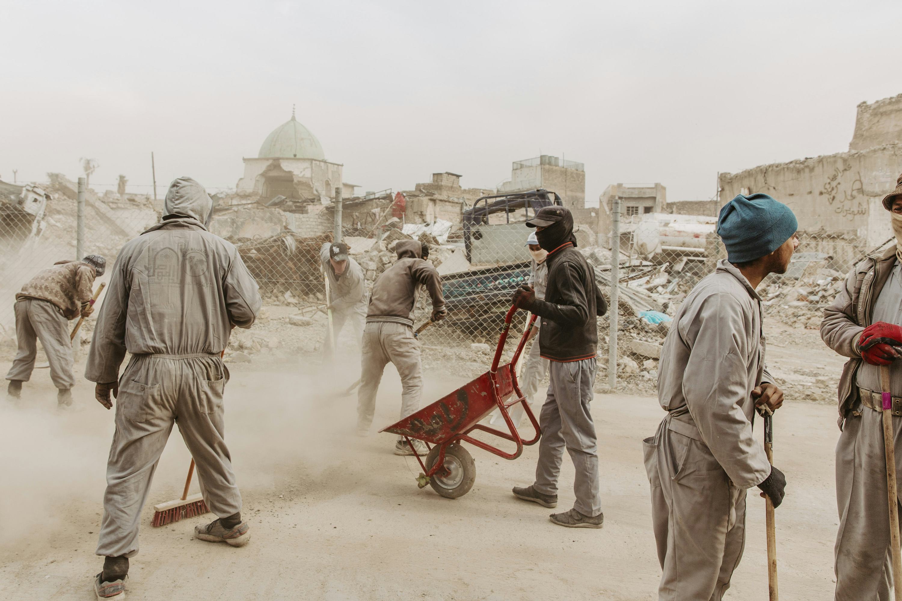 Image of various men in grey boilersuits cleaning up a dusty road, showing a red wheelbarrow in the middle and destroyed buildings in the background.