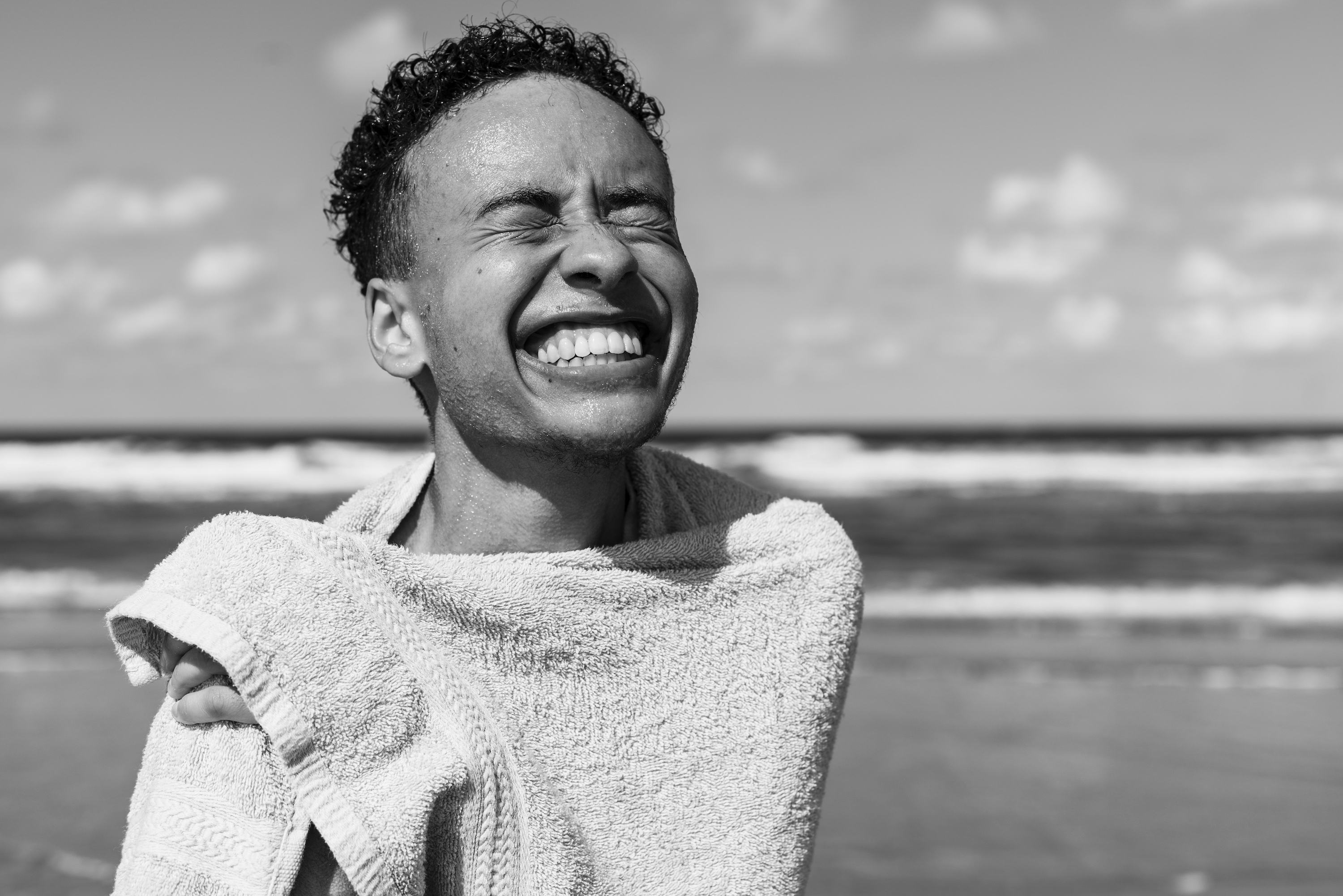 A black-and-white portrait of the photographer, Marvel Harris, on the beach, smiling brightly with his eyes closed, holding a towel around him.