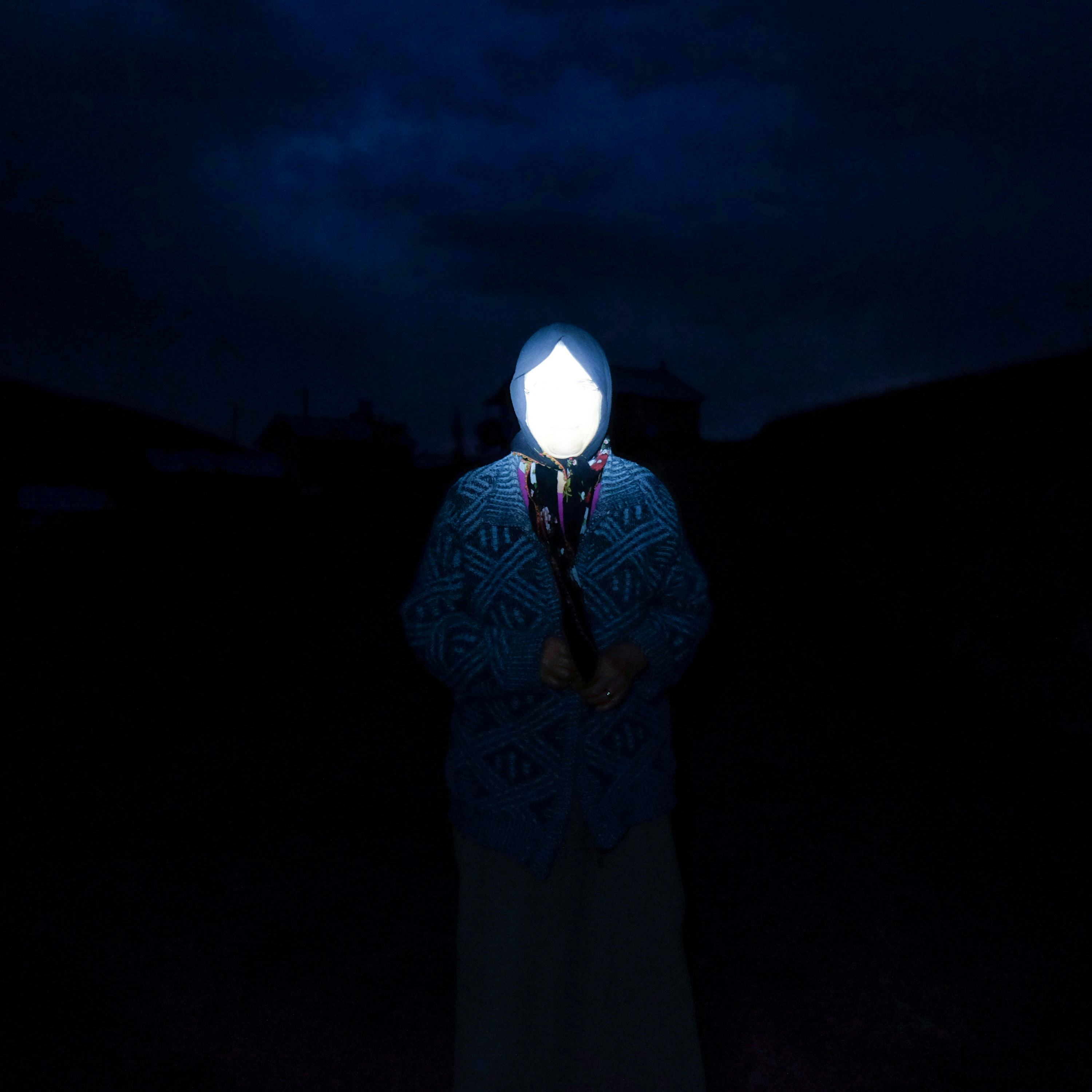 Portrait of a woman in the Kushmer Highlands at night, wearing a head scarf. Her face is invisible due to the flash of the camera ©Cansu Yıldıran