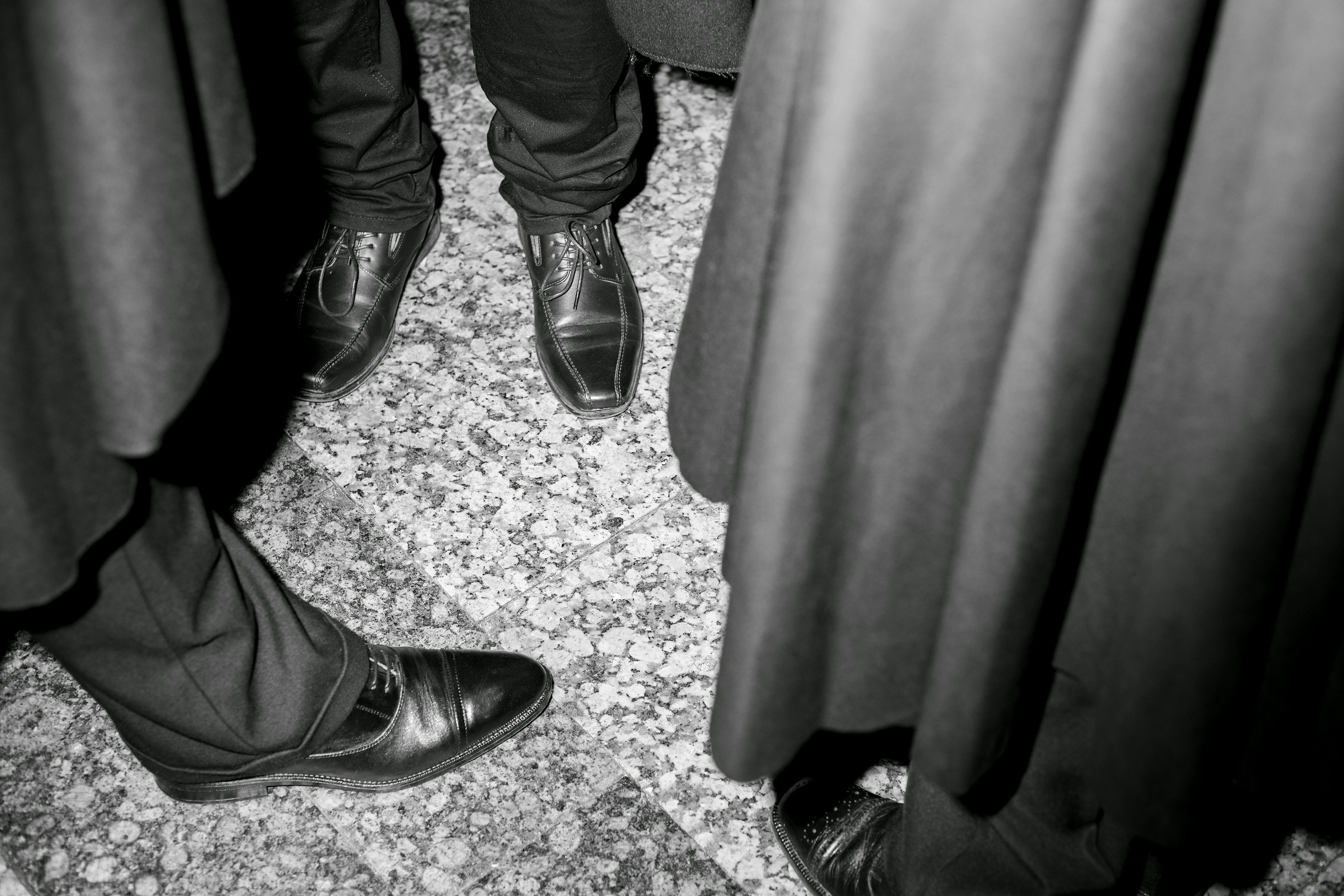 Black and white close-up shot of several pairs of feet, wearing fancy shoes, standing on a marble floor. © Bebe Blanco Agterberg