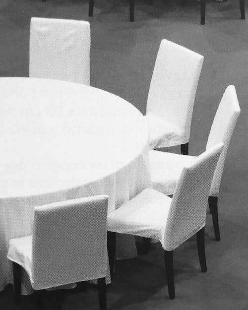 Black and white image showing a close-up of five chairs and a table, covered in white linen. © Eleonora Agostini