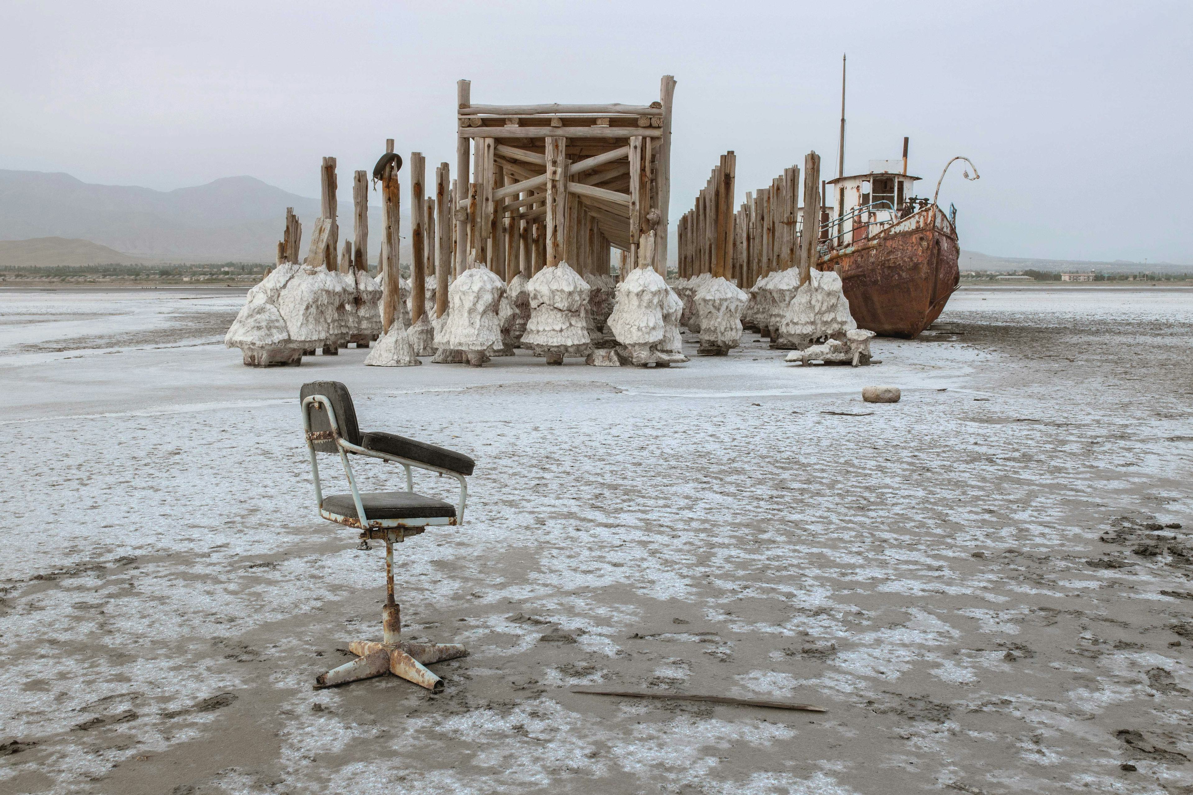 Photo of a worn down office chair on the floor of a dried-up salt lake, remnants of salt stuck the the pillars of the pier in the background. © Solmaz Daryani