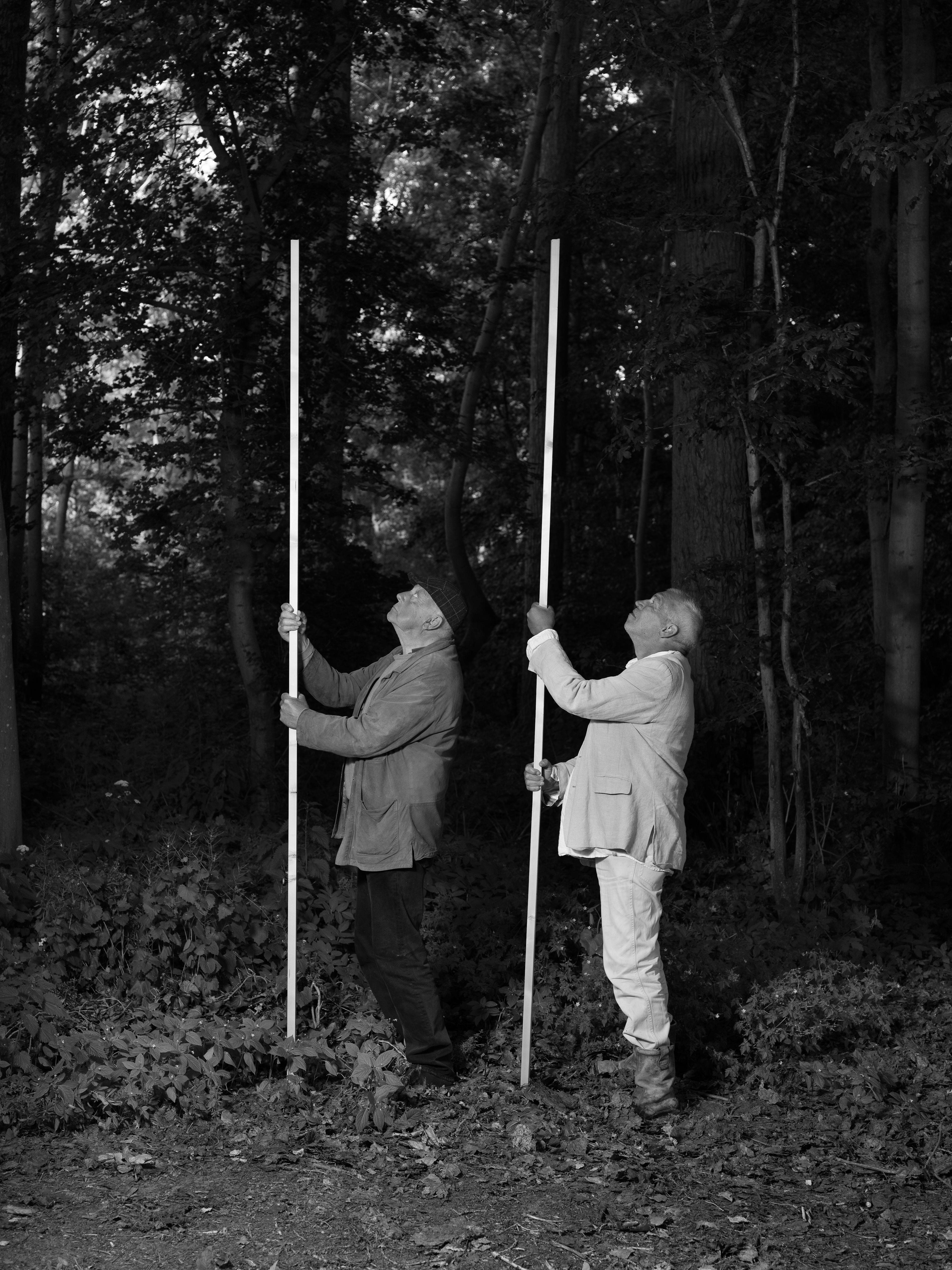 Black and white image of two men standing in a forest, both holding a long white vertical pole with both hands, whilst looking up. © Bebe Blanco Agterberg