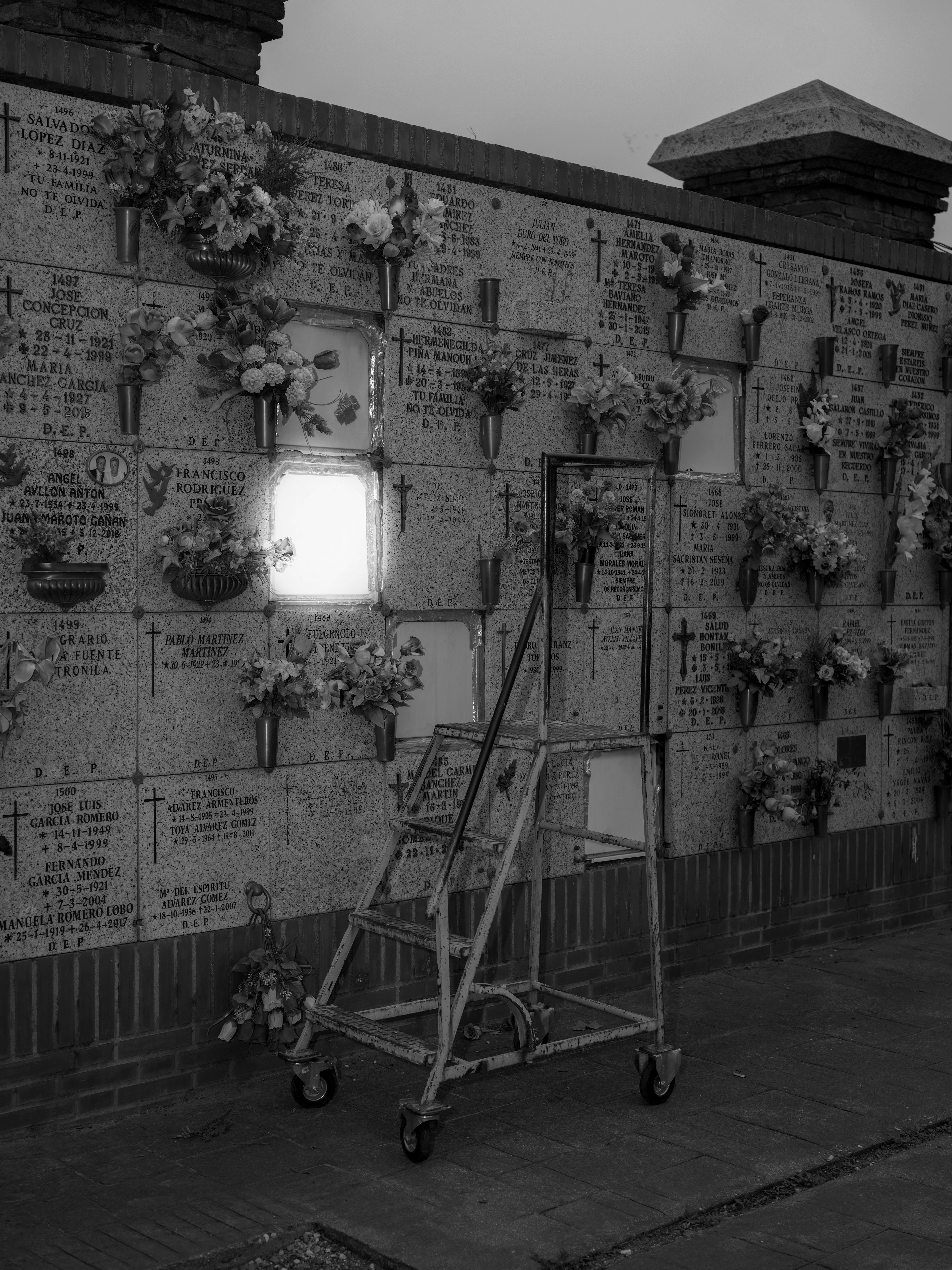 Black and white image of a wall with graves: showing epigraphs and flowers. In front is a pair of stairs. © Bebe Blanco Agterberg