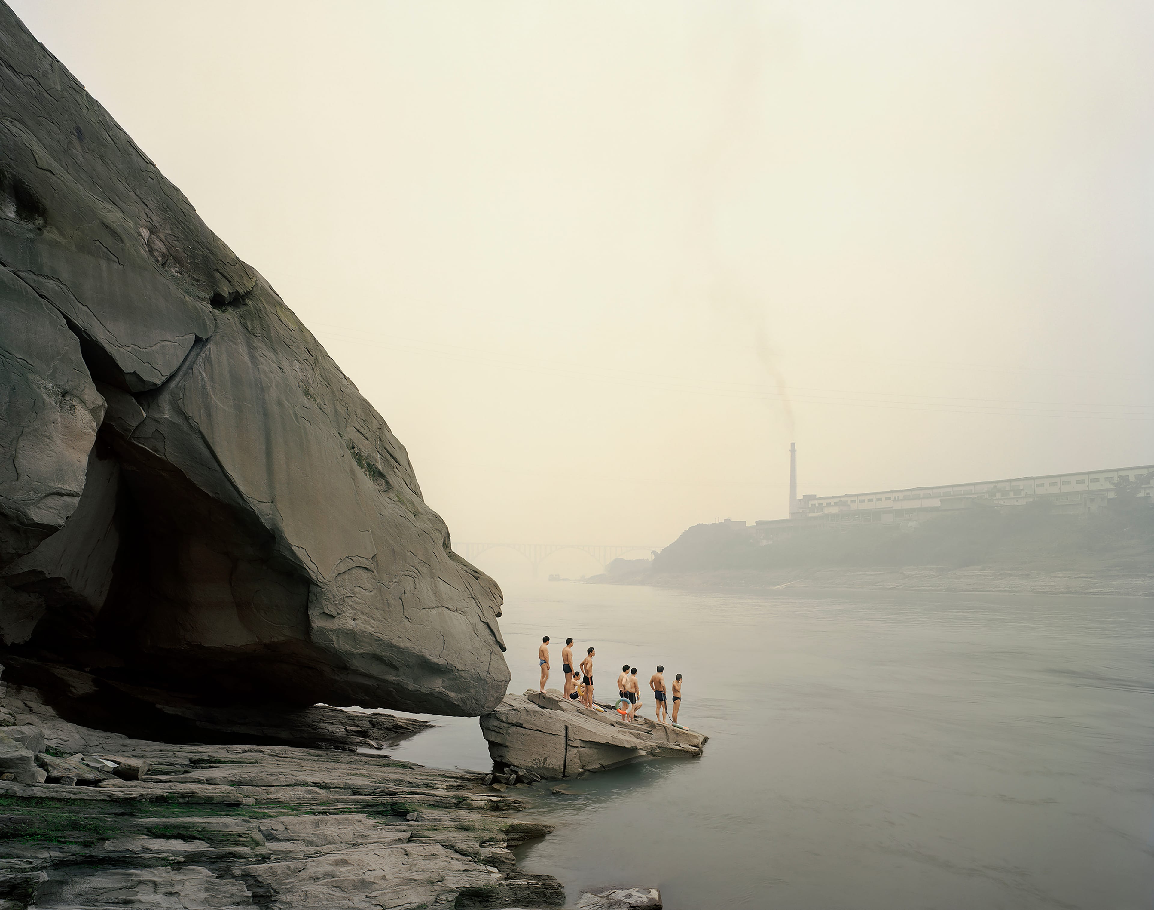 People stand on a rock by a foggy riverside, with a factory and bridge in the background. Smokestack emits smoke into the sky.