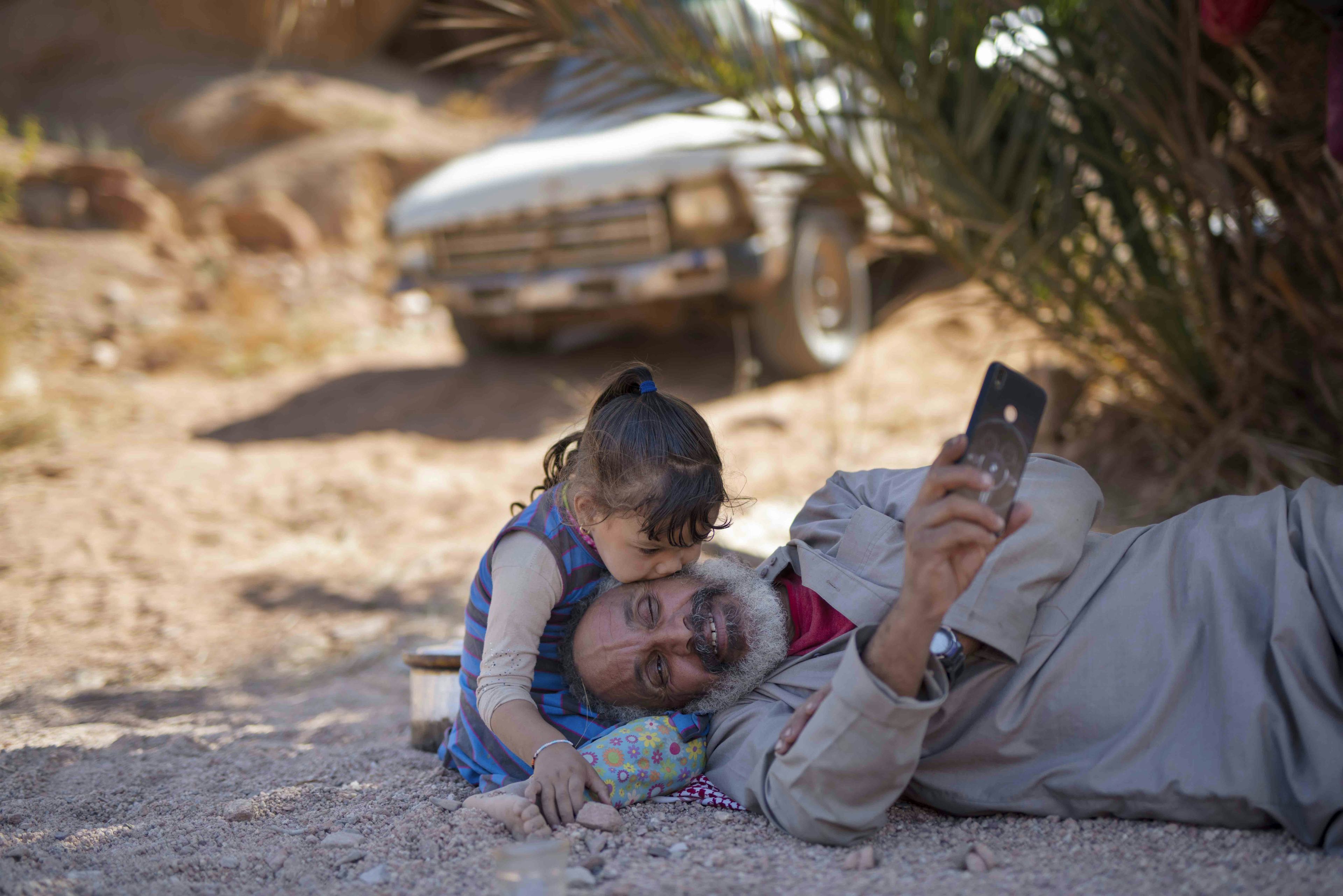 Photo of an older man resting on the floor, holding his phone, whilst a small girl sits besides him, kissing his cheek © Rehab Eldalil