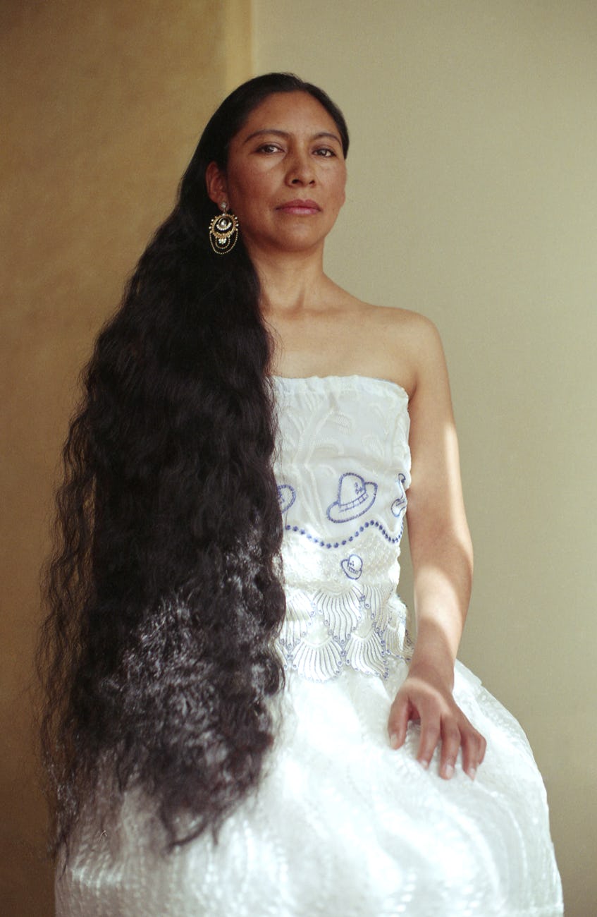 Portrait of a seated and solemn-looking woman wearing a white strapless dress and long brown curly hair over one shoulder © Marisol Mendez