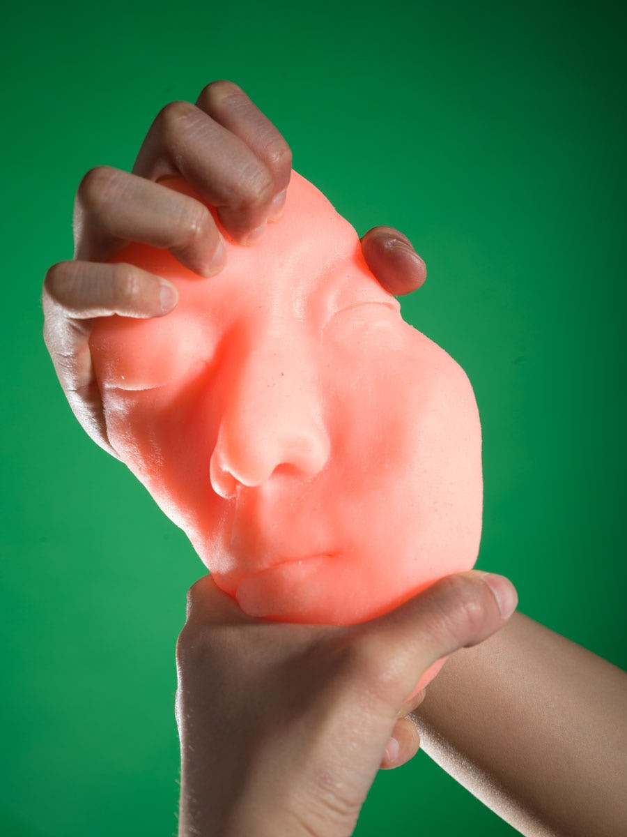 Pink silicon cast of the artist's face, in front of a green background, being twisted with two hands© Sheung Yiu